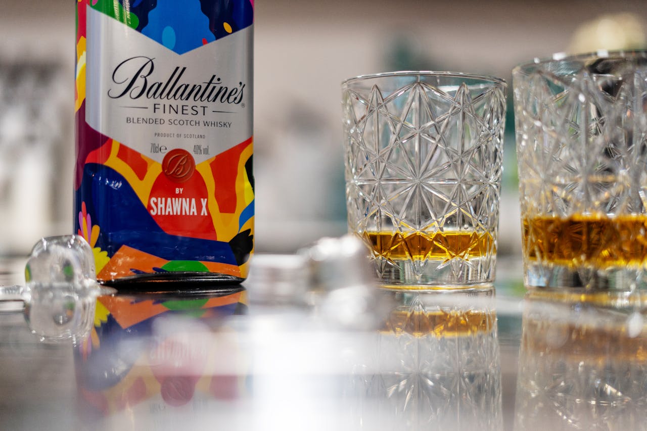 A vibrant Ballantines whisky bottle and glasses on a reflective surface with ice cubes.